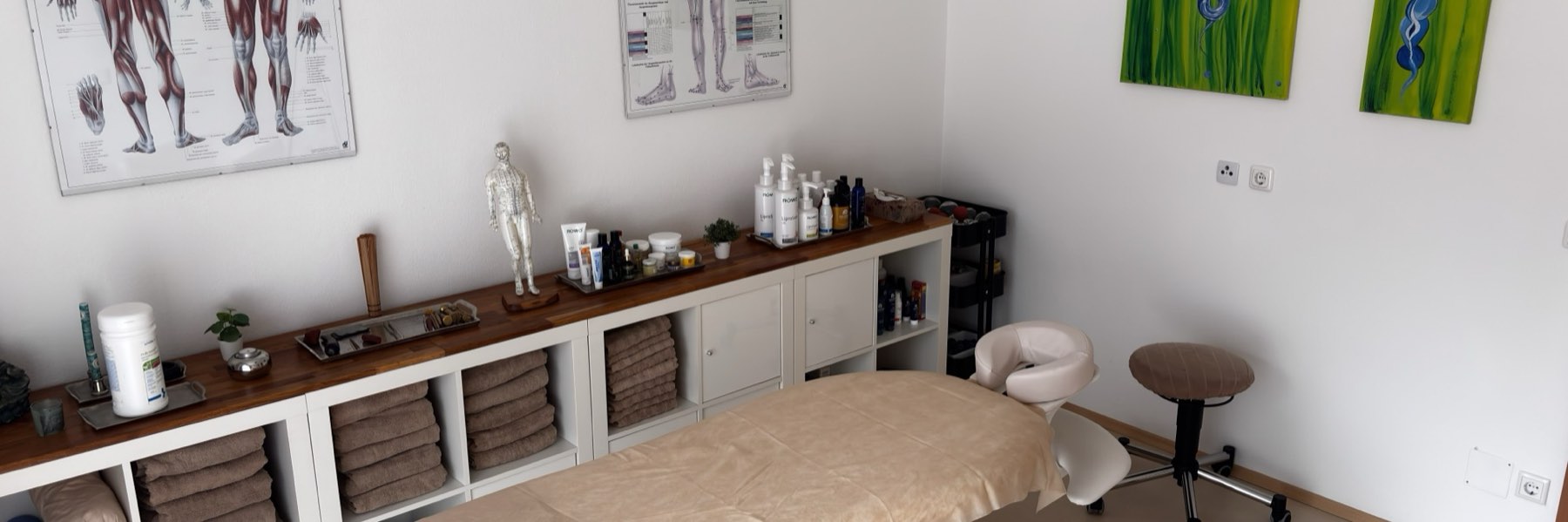 Der Behandlungsraum der Praxis: Massageliege, Regal mit Tüchern und Meridiantafeln an der Wand